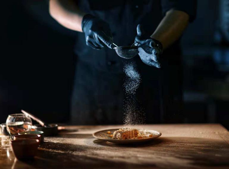 Chef dusting a dish at a dark kitchen pass